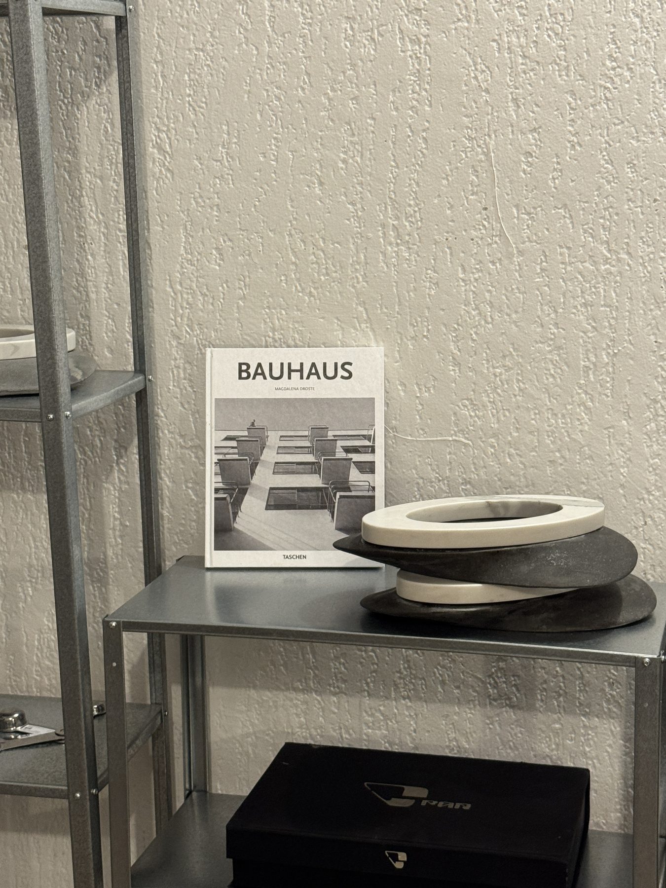 Pila de platos de mármol y concreto sobre un estante metálico, junto a un libro de Bauhaus y una caja negra de PAR